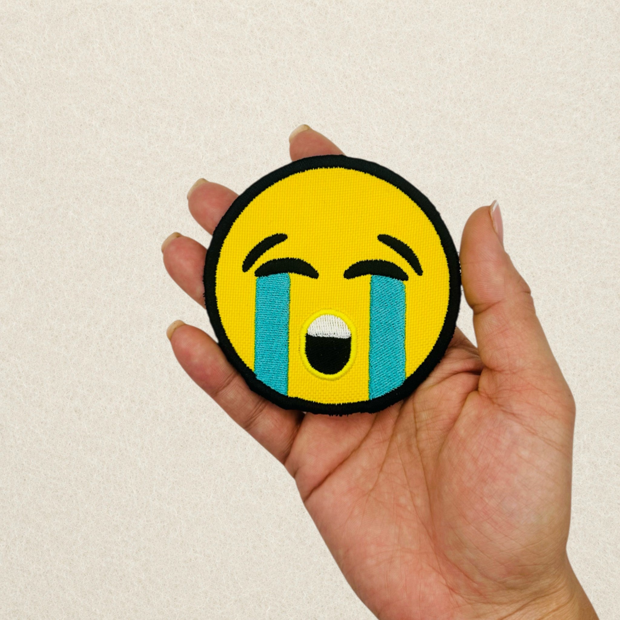 Crying Face Emoji embroidered patch shown in hand to demonstrate its actual size.