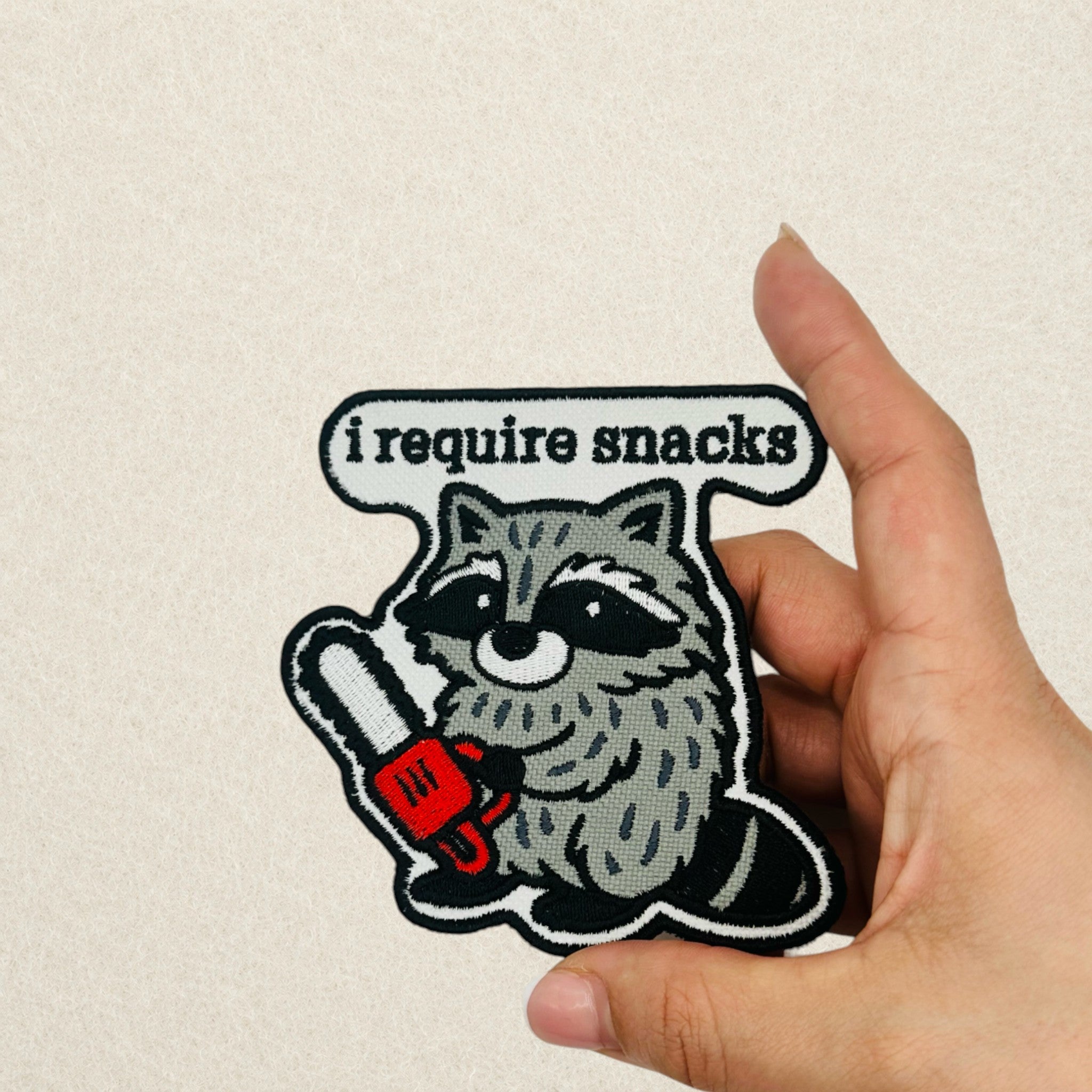 I Require Snacks Raccoon embroidered patch shown in hand to demonstrate its actual size.