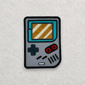 Retro Game Boy inspired embroidered patch displayed on a light grey felt background.